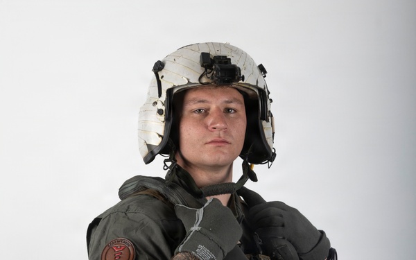 A young male in full military flight crew gear poses against a plain white/grey studio background. He is wearing a flight suit, survival vest, flight helmet with night vision mount, and tactical gloves. The image appears to be a professional portrait or documentation photograph.