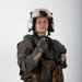 A young male in full military flight crew gear poses against a plain white/grey studio background. He is wearing a flight suit, survival vest, flight helmet with night vision mount, and tactical gloves. The image appears to be a professional portrait or documentation photograph.