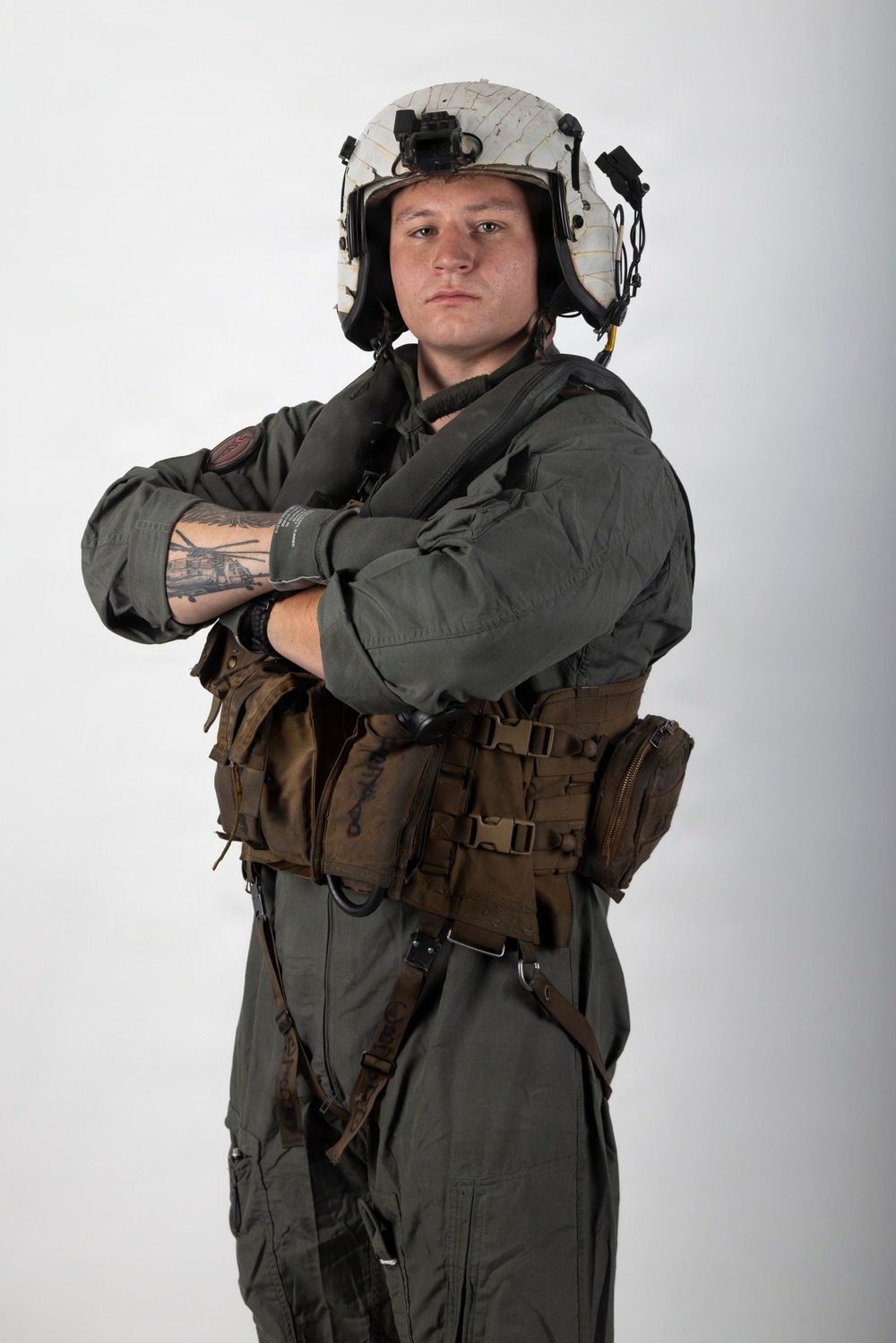 A military aviator poses against a plain white/light gray studio background with arms crossed. The individual is wearing full flight gear including a white flight helmet with mounted equipment, an olive drab flight suit, and a tactical survival vest. The portrait-style photograph appears to be an official or professional military photo.