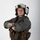 MCAS Marines pose for content 2026 A military aviator poses against a plain white/light gray studio background with arms crossed. The individual is wearing full flight gear including a white flight helmet with mounted equipment, an olive drab flight suit, and a tactical survival vest. The portrait-style photograph appears to be an official or professional military photo.