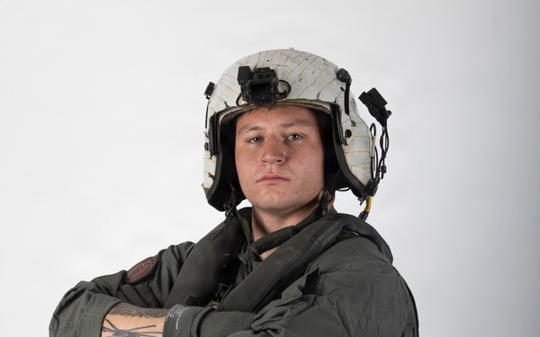 A military aviator poses against a plain white/light gray studio background with arms crossed. The individual is wearing full flight gear including a white flight helmet with mounted equipment, an olive drab flight suit, and a tactical survival vest. The portrait-style photograph appears to be an official or professional military photo.
