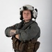 A military aviator poses against a plain white/light gray studio background with arms crossed. The individual is wearing full flight gear including a white flight helmet with mounted equipment, an olive drab flight suit, and a tactical survival vest. The portrait-style photograph appears to be an official or professional military photo.