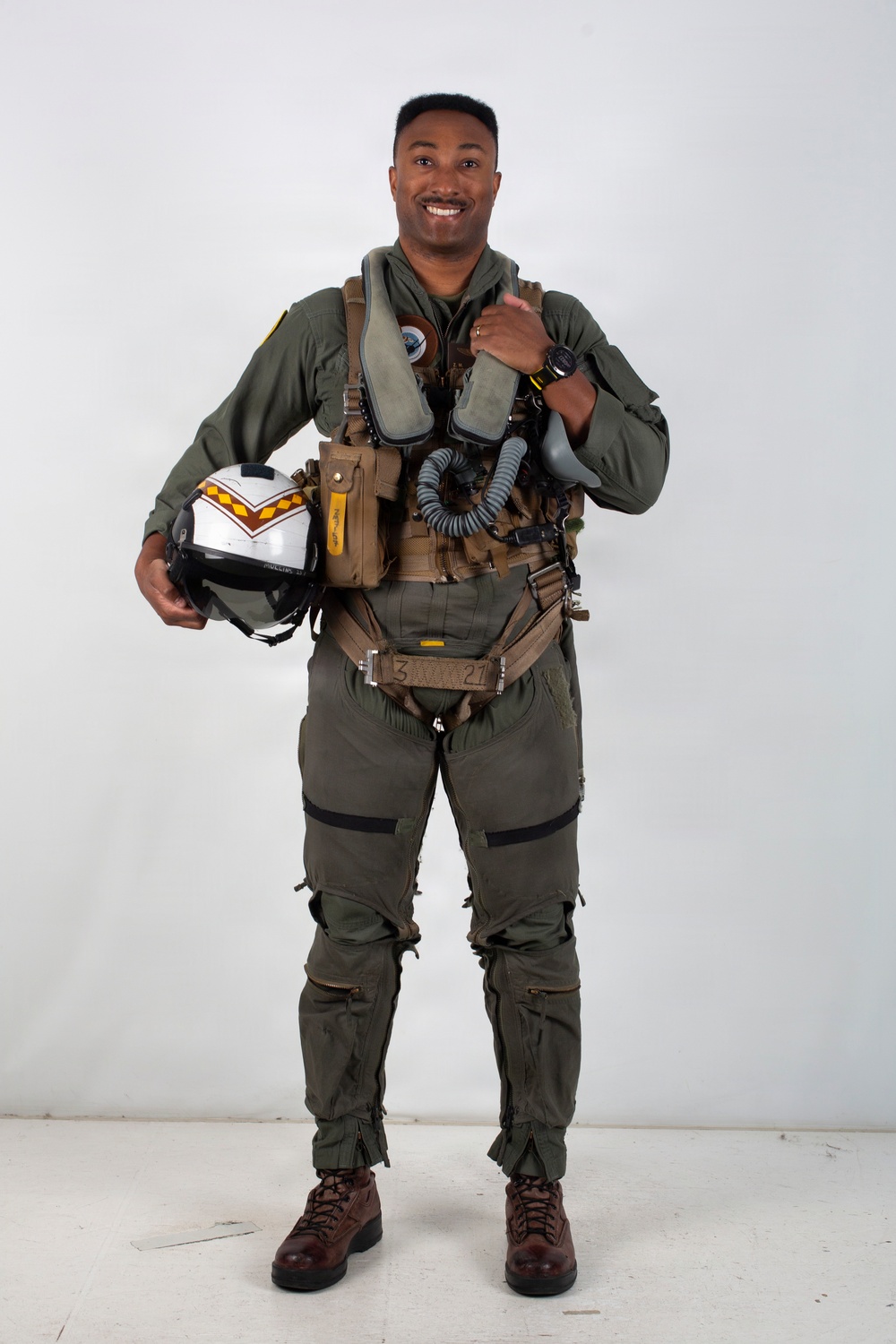 A military aviator poses in full flight gear against a plain white/light gray studio background. He is smiling and holding a flight helmet in one hand while giving a thumbs-up gesture with the other. The subject is dressed in a complete fighter pilot ensemble including a flight suit, harness, G-suit, survival vest, and oxygen mask assembly.