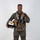 MCAS Marines pose for content 2026 A military aviator poses in full flight gear against a plain white/light gray studio background. He is smiling and holding a flight helmet in one hand while giving a thumbs-up gesture with the other. The subject is dressed in a complete fighter pilot ensemble including a flight suit, harness, G-suit, survival vest, and oxygen mask assembly.