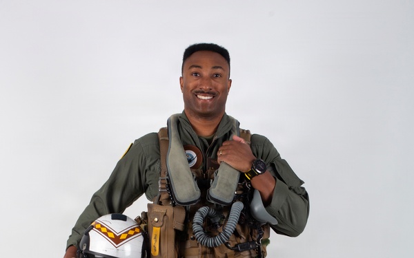 A military aviator poses in full flight gear against a plain white/light gray studio background. He is smiling and holding a flight helmet in one hand while giving a thumbs-up gesture with the other. The subject is dressed in a complete fighter pilot ensemble including a flight suit, harness, G-suit, survival vest, and oxygen mask assembly.