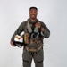A military aviator poses in full flight gear against a plain white/light gray studio background. He is smiling and holding a flight helmet in one hand while giving a thumbs-up gesture with the other. The subject is dressed in a complete fighter pilot ensemble including a flight suit, harness, G-suit, survival vest, and oxygen mask assembly.