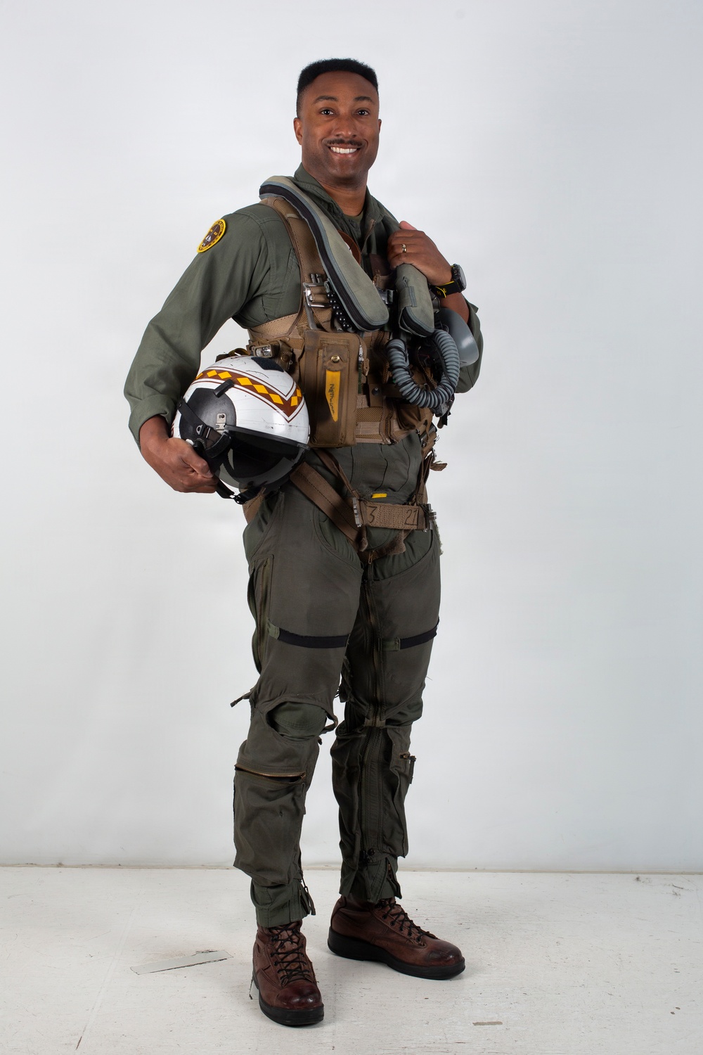 A military aviator poses in full flight gear against a plain white/light gray studio backdrop. He is smiling and holding a flight helmet with distinctive yellow and red chevron markings on a white shell. The image appears to be an official or professional portrait photograph.