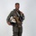 MCAS Marines pose for content 2026 A military aviator poses in full flight gear against a plain white/light gray studio backdrop. He is smiling and holding a flight helmet with distinctive yellow and red chevron markings on a white shell. The image appears to be an official or professional portrait photograph.