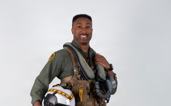 A military aviator poses in full flight gear against a plain white/light gray studio backdrop. He is smiling and holding a flight helmet with distinctive yellow and red chevron markings on a white shell. The image appears to be an official or professional portrait photograph.
