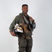A military aviator poses in full flight gear against a plain white/light gray studio backdrop. He is smiling and holding a flight helmet with distinctive yellow and red chevron markings on a white shell. The image appears to be an official or professional portrait photograph.