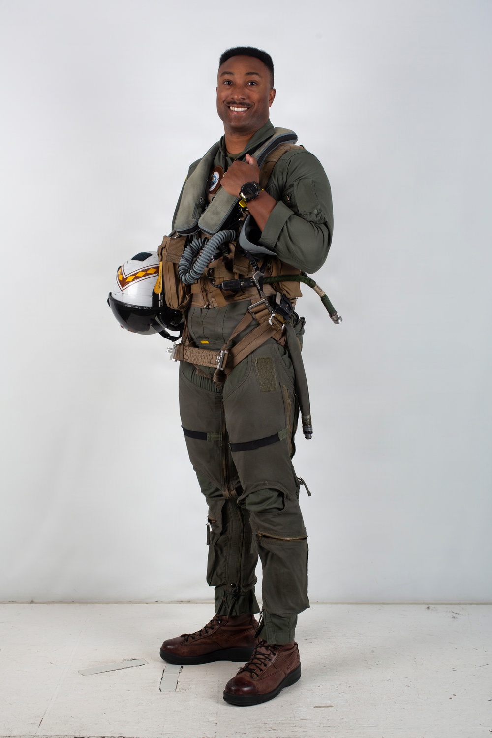 A military aviator stands in a full-body portrait pose against a plain light gray background. He is wearing a complete flight suit ensemble including a G-suit, survival vest, and harness, and is holding a flight helmet. He is smiling and appears to be posing for an official or promotional photograph.