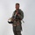MCAS Marines pose for content 2026 A military aviator stands in a full-body portrait pose against a plain light gray background. He is wearing a complete flight suit ensemble including a G-suit, survival vest, and harness, and is holding a flight helmet. He is smiling and appears to be posing for an official or promotional photograph.