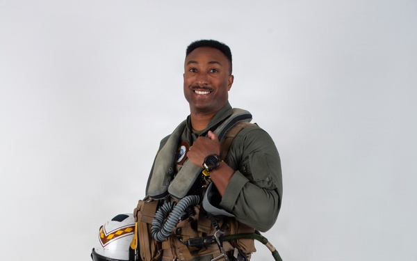A military aviator stands in a full-body portrait pose against a plain light gray background. He is wearing a complete flight suit ensemble including a G-suit, survival vest, and harness, and is holding a flight helmet. He is smiling and appears to be posing for an official or promotional photograph.