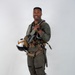 A military aviator stands in a full-body portrait pose against a plain light gray background. He is wearing a complete flight suit ensemble including a G-suit, survival vest, and harness, and is holding a flight helmet. He is smiling and appears to be posing for an official or promotional photograph.