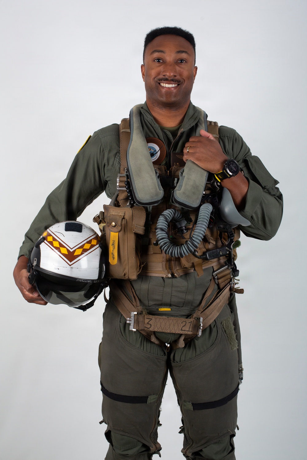A military aviator poses for a formal portrait against a neutral gray background, wearing a full flight suit and survival gear. He is smiling and holding a white flight helmet with brown and yellow chevron markings. His equipment suggests he is a jet fighter or tactical aircraft pilot.