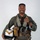 MCAS Marines pose for content 2026 A military aviator poses for a formal portrait against a neutral gray background, wearing a full flight suit and survival gear. He is smiling and holding a white flight helmet with brown and yellow chevron markings. His equipment suggests he is a jet fighter or tactical aircraft pilot.