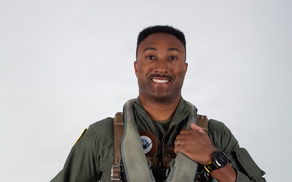 A military aviator poses for a formal portrait against a neutral gray background, wearing a full flight suit and survival gear. He is smiling and holding a white flight helmet with brown and yellow chevron markings. His equipment suggests he is a jet fighter or tactical aircraft pilot.