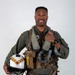 A military aviator poses for a formal portrait against a neutral gray background, wearing a full flight suit and survival gear. He is smiling and holding a white flight helmet with brown and yellow chevron markings. His equipment suggests he is a jet fighter or tactical aircraft pilot.
