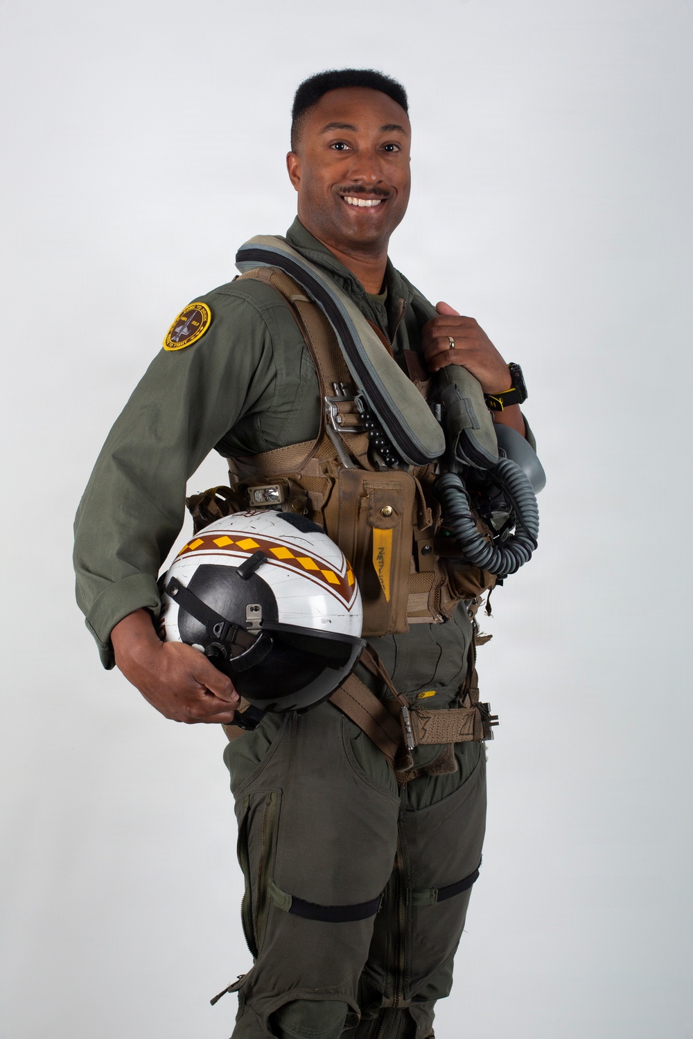 A military aviator poses in a professional portrait-style photograph against a neutral gray background. He is wearing a full flight suit ensemble and holding a decorated flight helmet, smiling confidently at the camera. The image appears to be an official or promotional military photograph.