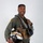 MCAS Marines pose for content 2026 A military aviator poses in a professional portrait-style photograph against a neutral gray background. He is wearing a full flight suit ensemble and holding a decorated flight helmet, smiling confidently at the camera. The image appears to be an official or promotional military photograph.