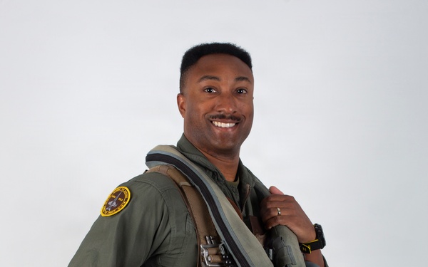 A military aviator poses in a professional portrait-style photograph against a neutral gray background. He is wearing a full flight suit ensemble and holding a decorated flight helmet, smiling confidently at the camera. The image appears to be an official or promotional military photograph.
