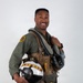 A military aviator poses in a professional portrait-style photograph against a neutral gray background. He is wearing a full flight suit ensemble and holding a decorated flight helmet, smiling confidently at the camera. The image appears to be an official or promotional military photograph.