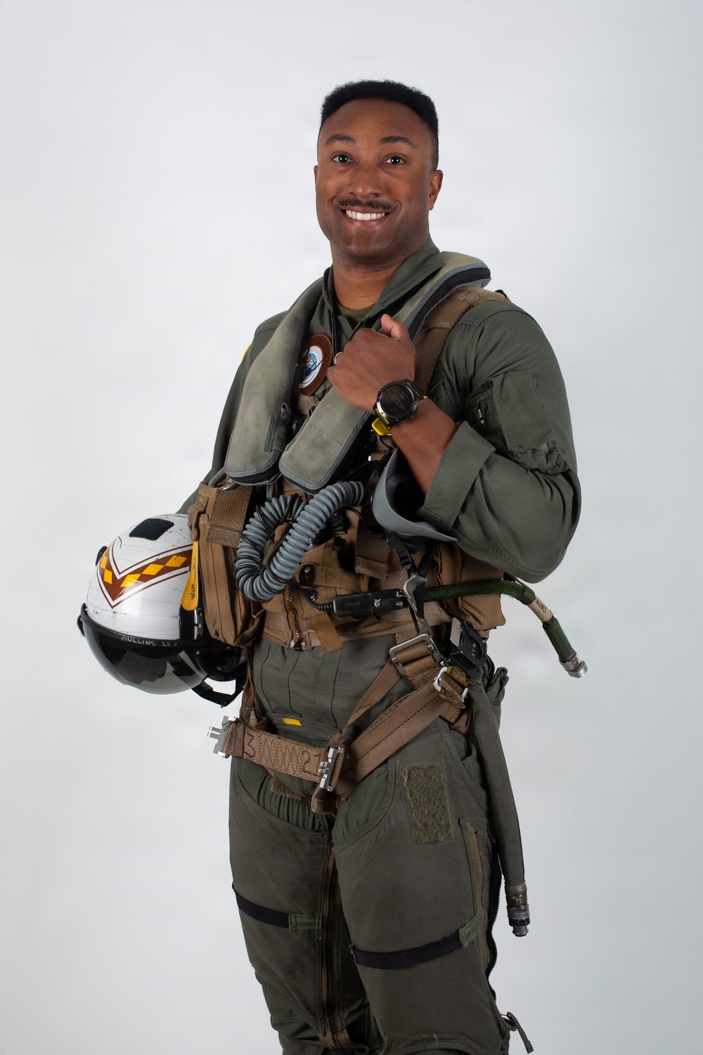 A military aviator poses in full flight gear against a neutral light gray background in what appears to be an official portrait photograph. The subject is smiling and holding a flight helmet under one arm while wearing a complete ensemble of aviation equipment. The image is professionally lit and composed.