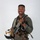 MCAS Marines pose for content 2026 A military aviator poses in full flight gear against a neutral light gray background in what appears to be an official portrait photograph. The subject is smiling and holding a flight helmet under one arm while wearing a complete ensemble of aviation equipment. The image is professionally lit and composed.