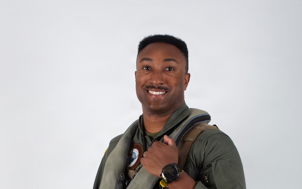 A military aviator poses in full flight gear against a neutral light gray background in what appears to be an official portrait photograph. The subject is smiling and holding a flight helmet under one arm while wearing a complete ensemble of aviation equipment. The image is professionally lit and composed.