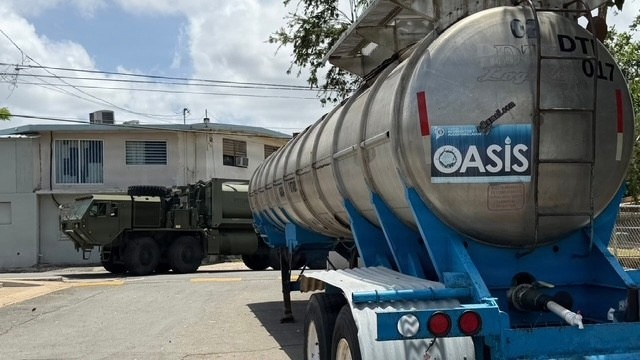 Military Vehicle and Oasis Water Tanker Operate Near Residential Building During Water Distribution Mission