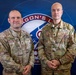 Two U.S. Army soldiers in OCP (Operational Camouflage Pattern) uniforms pose for a formal photo in front of a blue backdrop featuring a military unit insignia partially reading 'AGON'S L' (likely 'Dragon's Lair' or similar). The soldier on the left is identified by name tape as 'POTTS' and the soldier on the right as 'KIMBIRSKYI', both wearing U.S. Army name tapes. The image appears to be an official military portrait or recognition photo.