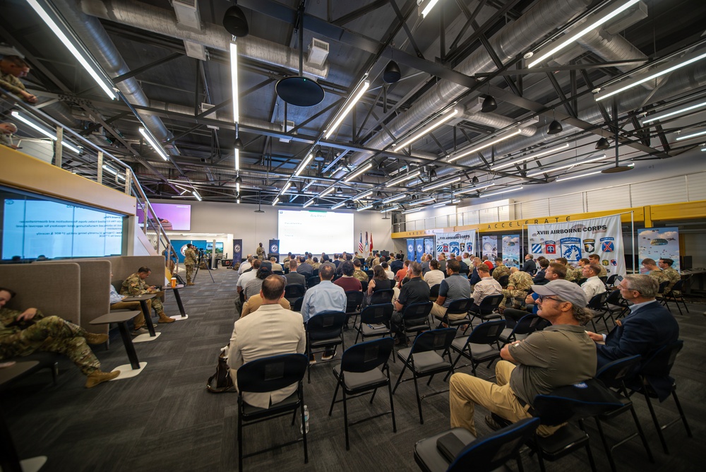 A large briefing or presentation event is taking place in a modern industrial-style facility associated with the XVIII Airborne Corps. The audience consists of a mix of military personnel in camouflage uniforms and civilians seated in rows of folding chairs facing a projection screen at the front. Banners and signage on the right wall display XVIII Airborne Corps branding and unit patches, with the word 'ACCELERATE' prominently visible.