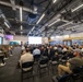 A large briefing or presentation event is taking place in a modern industrial-style facility associated with the XVIII Airborne Corps. The audience consists of a mix of military personnel in camouflage uniforms and civilians seated in rows of folding chairs facing a projection screen at the front. Banners and signage on the right wall display XVIII Airborne Corps branding and unit patches, with the word 'ACCELERATE' prominently visible.