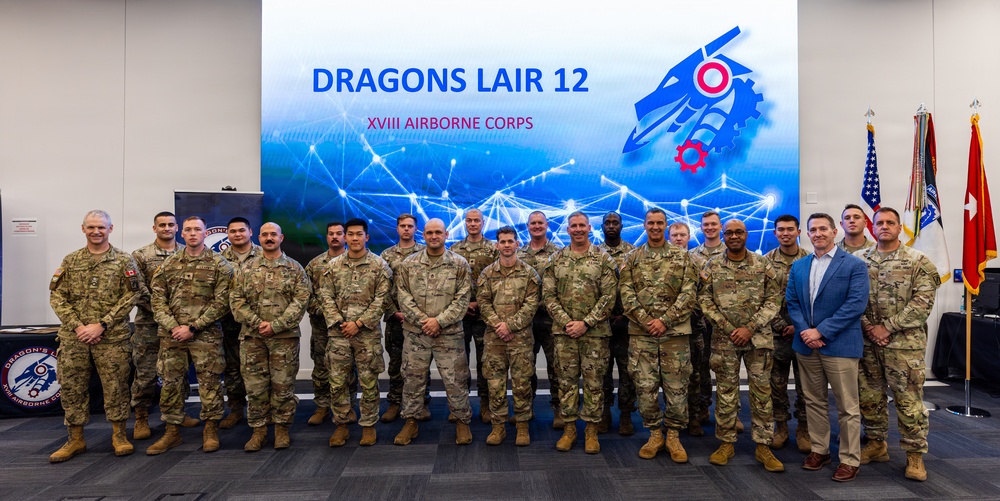 A group photo of approximately 22 military personnel and one civilian posing in front of a large display screen reading 'Dragons Lair 12 - XVIII Airborne Corps.' The participants are predominantly wearing U.S. Army OCP (Operational Camouflage Pattern) uniforms, with one individual in civilian business attire. The setting appears to be an indoor conference or event facility with military flags visible on the right side.