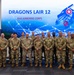 A group photo of approximately 22 military personnel and one civilian posing in front of a large display screen reading 'Dragons Lair 12 - XVIII Airborne Corps.' The participants are predominantly wearing U.S. Army OCP (Operational Camouflage Pattern) uniforms, with one individual in civilian business attire. The setting appears to be an indoor conference or event facility with military flags visible on the right side.