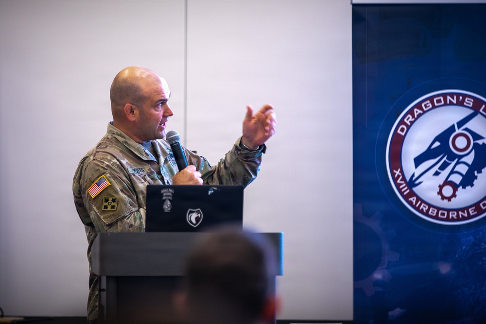 A U.S. Army soldier in camouflage uniform is delivering a presentation at a podium with a microphone, gesturing with his right hand. A laptop is visible on the podium. Behind him is a banner displaying the 'Dragon's Den - XVIII Airborne Corps' logo.