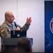 A U.S. Army soldier in camouflage uniform is delivering a presentation at a podium with a microphone, gesturing with his right hand. A laptop is visible on the podium. Behind him is a banner displaying the 'Dragon's Den - XVIII Airborne Corps' logo.