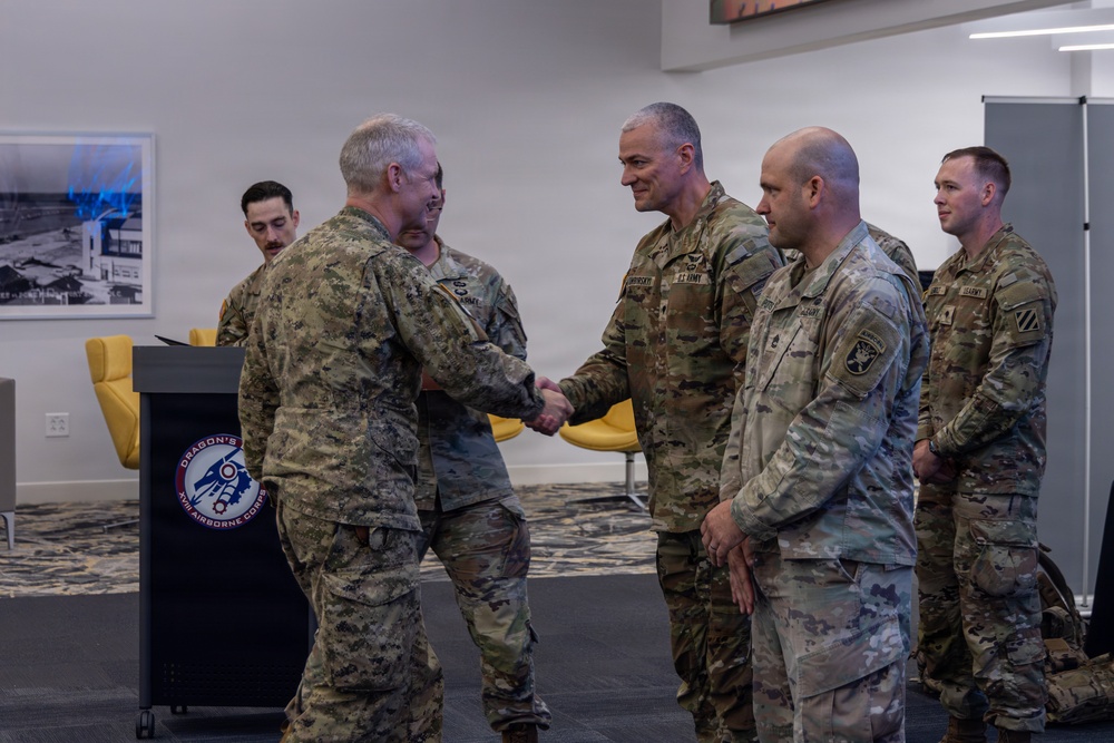 A formal military greeting ceremony is taking place indoors, likely at a U.S. Army installation. A senior officer in a distinctive camouflage uniform is shaking hands with another officer while additional soldiers stand in a receiving line. A podium bearing the XVIII Airborne Corps 'Dragon's' insignia is visible in the foreground.