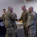 A formal military greeting ceremony is taking place indoors, likely at a U.S. Army installation. A senior officer in a distinctive camouflage uniform is shaking hands with another officer while additional soldiers stand in a receiving line. A podium bearing the XVIII Airborne Corps 'Dragon's' insignia is visible in the foreground.