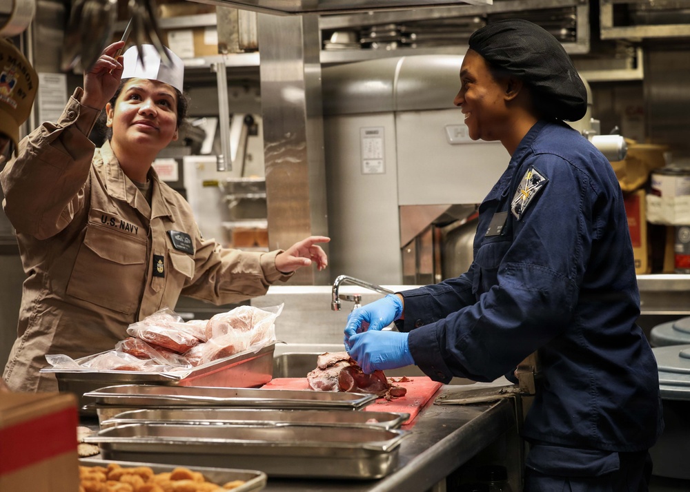 Two U.S. Navy personnel are working in a military galley (shipboard kitchen). The person on the left wears a tan U.S. Navy uniform with a white paper chef's hat and holds a knife, while the person on the right wears a blue coverall uniform with a black hair net and blue latex gloves, cutting meat on a red cutting board. Both individuals appear to be engaged in conversation while preparing food.