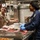 Two U.S. Navy personnel are working in a military galley (shipboard kitchen). The person on the left wears a tan U.S. Navy uniform with a white paper chef's hat and holds a knife, while the person on the right wears a blue coverall uniform with a black hair net and blue latex gloves, cutting meat on a red cutting board. Both individuals appear to be engaged in conversation while preparing food.