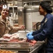 Two U.S. Navy personnel are working in a military galley (shipboard kitchen). The person on the left wears a tan U.S. Navy uniform with a white paper chef's hat and holds a knife, while the person on the right wears a blue coverall uniform with a black hair net and blue latex gloves, cutting meat on a red cutting board. Both individuals appear to be engaged in conversation while preparing food.