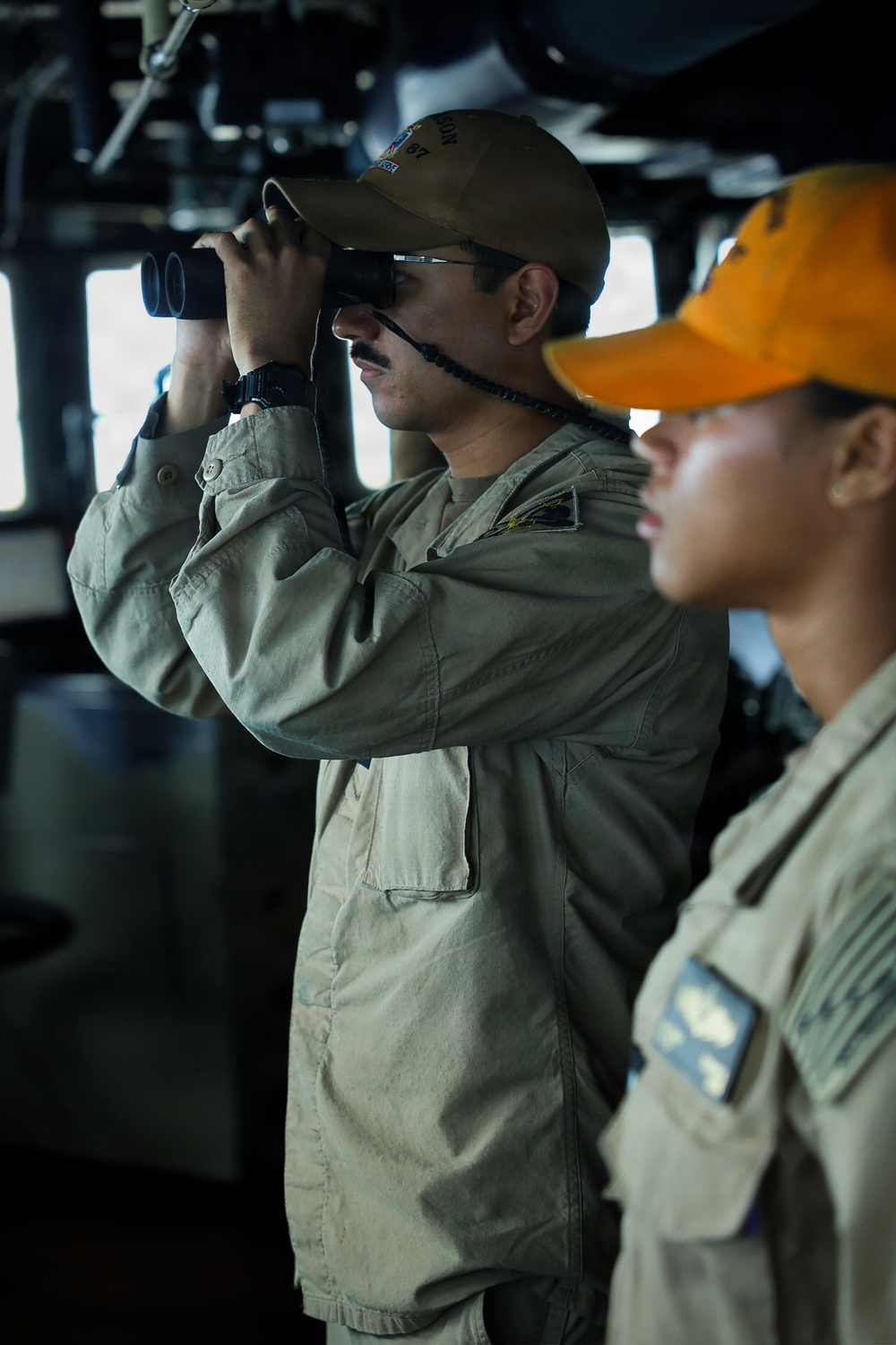 Two military personnel are stationed on what appears to be a ship's bridge or command deck. The individual in the foreground is actively scanning through binoculars while wearing a tan/olive uniform and a brown cap with partial text visible. A second person in the background wears an orange cap and similar uniform with a visible shoulder patch.