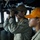 Two military personnel are stationed on what appears to be a ship's bridge or command deck. The individual in the foreground is actively scanning through binoculars while wearing a tan/olive uniform and a brown cap with partial text visible. A second person in the background wears an orange cap and similar uniform with a visible shoulder patch.