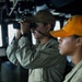 Two military personnel are stationed on what appears to be a ship's bridge or command deck. The individual in the foreground is actively scanning through binoculars while wearing a tan/olive uniform and a brown cap with partial text visible. A second person in the background wears an orange cap and similar uniform with a visible shoulder patch.