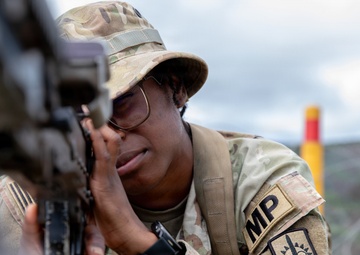 A U.S. Army Military Police (MP) soldier is in a prone firing position, aiming a rifle downrange at what appears to be a weapons qualification range. The soldier is wearing MultiCam (OCP) camouflage uniform with an 'MP' tab and a unit patch visible on the left sleeve, along with a boonie hat and glasses. The image is taken from a low angle, close to the weapon, giving a ground-level perspective of the training exercise.
