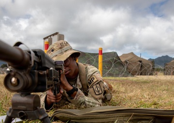 A U.S. Army Military Police soldier in OCP (Operational Camouflage Pattern) uniform is in a prone firing position on a grass field, aiming down the sights of a weapon mounted on a bipod. The setting appears to be a military training range or forward operating base, identifiable by military tents, concertina wire, and range markers in the background. A rifle case is visible on the ground beside the soldier.