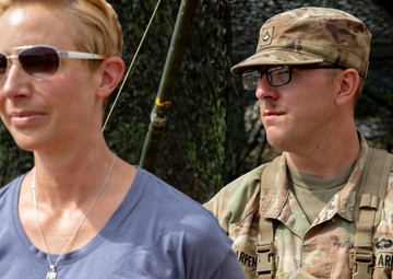 Two individuals are standing in front of a camouflage netting structure. One person is in civilian clothing wearing sunglasses and a necklace, while the other is a U.S. Army Military Police soldier in full OCP (Operational Camouflage Pattern) uniform holding a folder with documents. Both appear to be observing something off-camera.