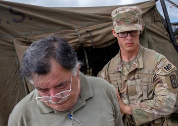 A U.S. Army Military Police (MP) soldier in full camouflage uniform is escorting or guiding a civilian man near a military tent marked 'U.S.' The soldier has his hand on the civilian's arm, suggesting a controlled escort situation. The setting appears to be an outdoor military field environment.