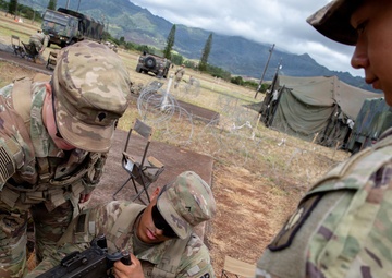 Three U.S. Army soldiers in OCP (Operational Camouflage Pattern) uniforms are gathered around a crew-served weapon, appearing to conduct training or maintenance on the firearm. One soldier wearing an 'MP' (Military Police) patch is kneeling and handling the weapon while another leans in to supervise or assist. The setting appears to be a field training environment with military tents, vehicles, and concertina wire visible in the background.
